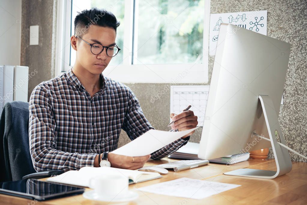 Young concentrated Asian man in checked shirt and glasses sitting at office desk and analyzing financial papers