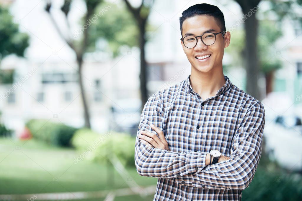 Waist-up portrait of attractive Asian guy in casual outfit and glasses standing outdoors with crossed hands and smiling at camera