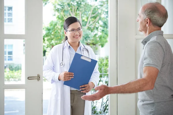 Elderly patient opening door to smiling Asian doctor with clipboard making visit