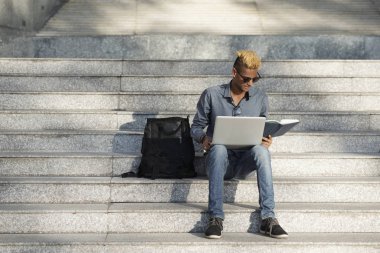 Basamaklarında oturan, bir kitap okuma ve dizüstü bilgisayar açık havada üzerinde çalışma Üniversitesi Öğrenci