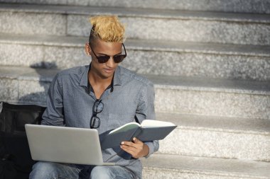 Bir kitap okuma ve yeni programlama dili öğrenirken dizüstü bilgisayarda çalışan genç siyah adam