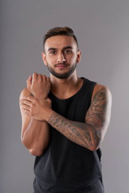 Handsome young bearded man with tattooed arm warming up wrists and hands looking at camera on gray background clipart