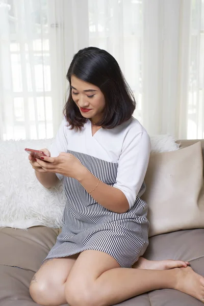 穿着漂亮衣服的亚洲女人微笑着坐在沙发上 在家的手机上打留言 — 图库照片