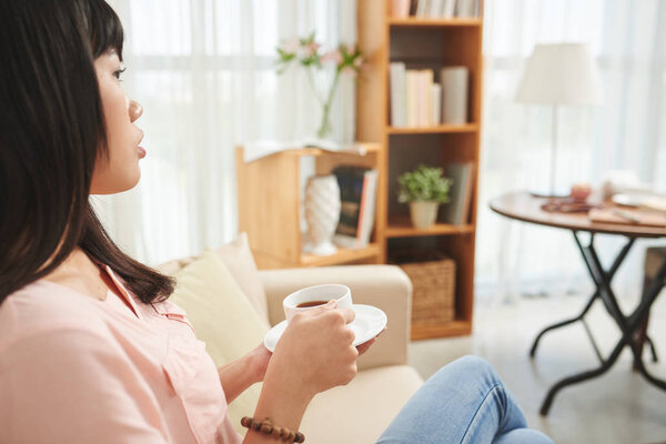 Pensive Vietnamese woman resting on sofa and drinking cup of black coffee 