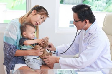Küçük bir çocuk annesi hastanede ile otururken incelenmesi için bir stetoskop kullanarak Asya erkek doktor