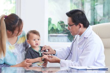 Küçük bir çocuk annesi hastanede ile otururken incelenmesi için bir stetoskop kullanarak Asya erkek doktor