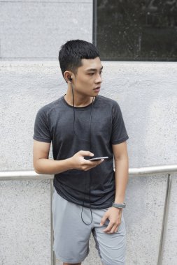 Ciddi genç Vietnamca adam açık havada duran ve onun smartphone cep telefonu ile müzik dinlemek 