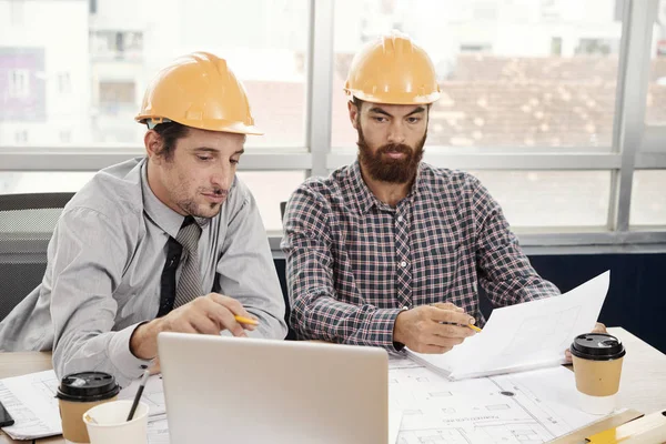 Seriöse Ingenieure Mit Harten Hüten Vergleichen Baupläne Auf Dem Laptop — Stockfoto