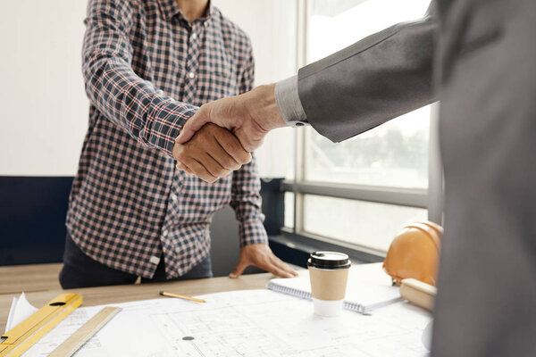 Business partners shaking hands over office table with blueprints on it