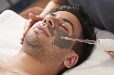 Genç yakışıklı adam gözleriyle rahatlatıcı kil maskesi uygulamak ve yordam Temizleme ultrason yapacağım güzellik uzmanı kapalı