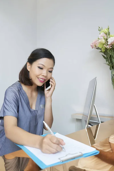 微笑的女性接待员在健康和美容院的接待处后面用手机交谈时 用文件做笔记 — 图库照片