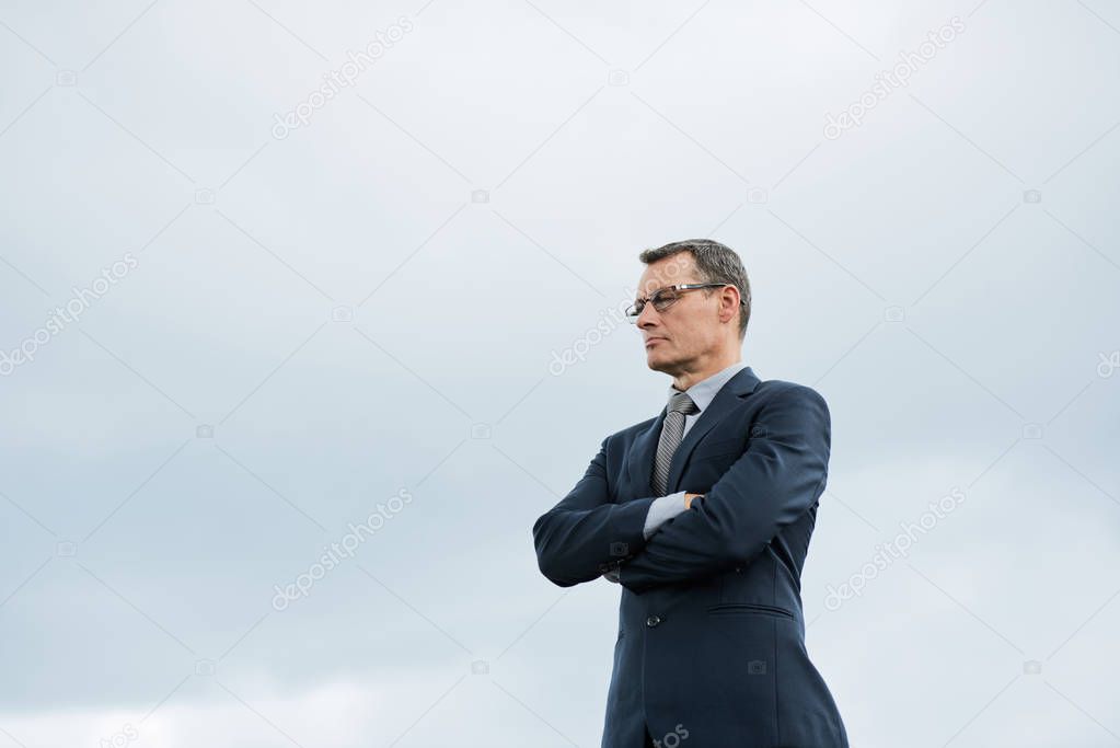 Caucasian businessman in suit against grey sky with arms crossed 