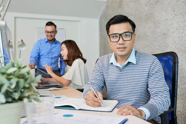 在现代办公室里 戴眼镜的休闲亚洲人坐在记事本上 同事在背景 — 图库照片