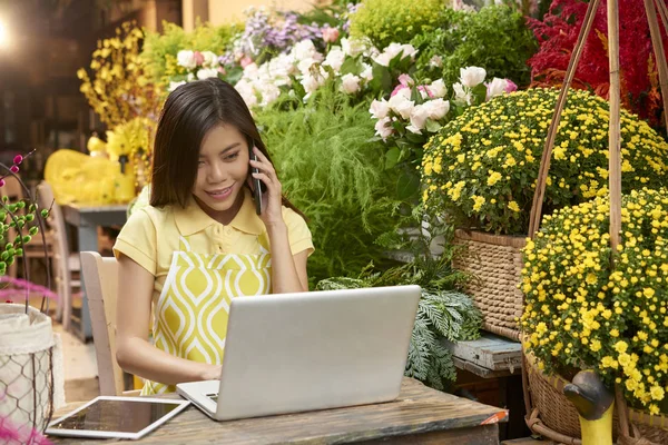 Pretty smiling young Asian woman making phone call to order flowers for her store