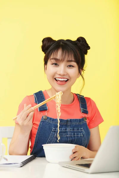 年轻的亚洲妇女用筷子从盒子里吃面条 在黄色背景的工作台上对着镜头微笑 — 图库照片