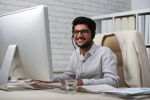 オフィスにあるコンピューターで作業するときにヘッドセットを着てメガネのハンサムな若いインドの起業家 — ストック写真