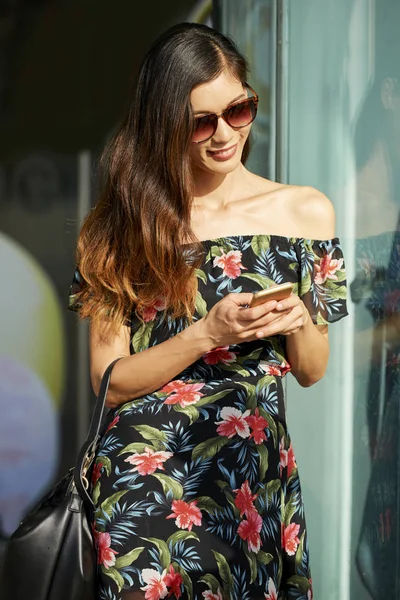 美丽的亚洲女性在花卉礼服微笑 并使用现代智能手机 而站在城市街道上的商店窗口附近的阳光明媚的日子 — 图库照片