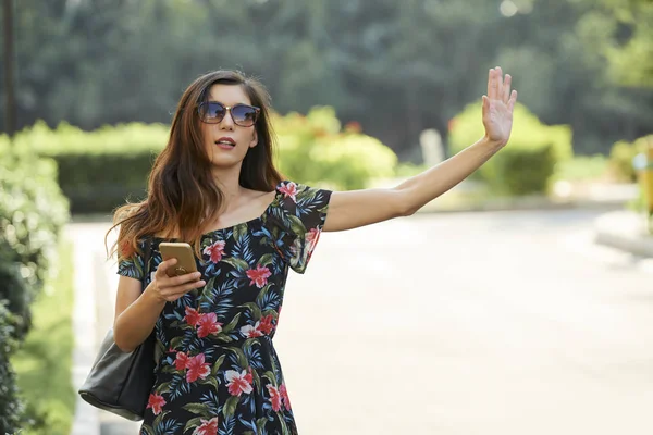 迷人的亚洲女性穿着时尚的服装 拿着智能手机 挥手致意 同时试图阻止公园附近的出租车 — 图库照片