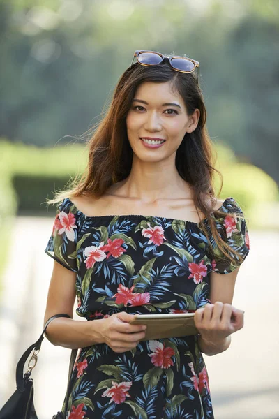 美丽的亚洲女人在花卉礼服举行平板电脑和看着相机与友好的脸的表情 而站在模糊的背景街道在阳光明媚的日子 — 图库照片