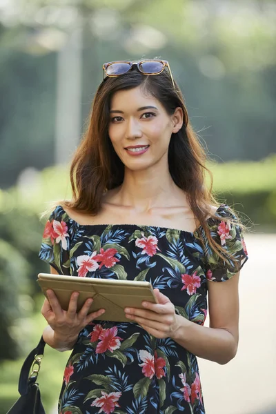 トレンディなドレスは笑みを浮かべて現代タブレットを保持し 晴れた日に公園の背景をぼかしの上に立ってながらカメラ目線でかわいいアジアの女性 — ストック写真