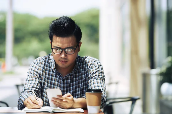 Geconcentreerde Jonge Aziatische Man Het Controleren Van Smartphone Het Opschrijven — Stockfoto