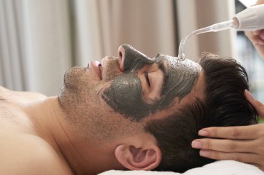 Yüzünde kil maskesi olan genç adam Spa salonunda Ultrasonik temizlik yapıyor