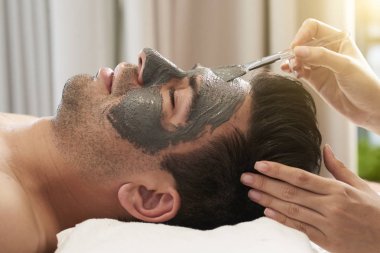 Genç adam yalan ve rahatlatıcı iken Kozmetolog Spa salonunda yüzünde kil maskesi uygulayarak