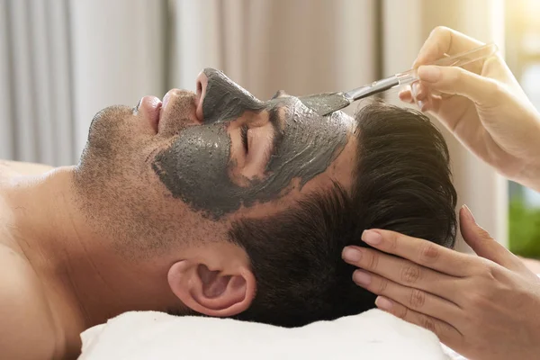 Jovem Deitado Relaxante Enquanto Cosmetologista Aplicando Máscara Barro Seu Rosto — Fotografia de Stock