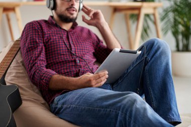 Casual giyim Tablet PC ile kanepede dinlenme genç adam, o kulaklık giyiyor ve Touchpad bir şey izliyor