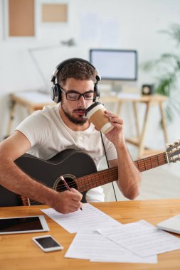 Stüdyo bir melodi oluştururken gitar ve içme kahve ile oturan genç müzisyen