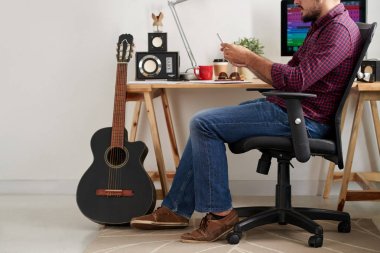 Bilgisayar ve gitar ile masaya yakın sandalyede oturan genç rahat adam ve ses stüdyosunda çalışırken onun Smartphone kullanarak
