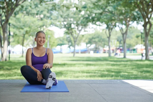 Laughing bald young Asian woman exercising on yoga mat in city park