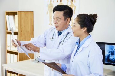 Üniformalı iki Asyalı tıp uzmanı, x-ray görüntüsünü işaret ediyor ve ofiste birlikte tartışıyorlar.
