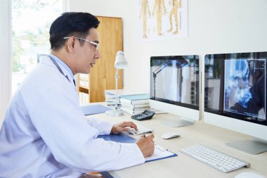 Gözlüklü Asyalı olgun nörolog bilgisayar monitöründe x-ray görüntüsünü inceliyor ve işyerinde çalışırken panoya notlar alıyor
