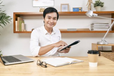 Ofis masasında tablet bilgisayar üzerinde çalışan pozitif genç Vietnamlı girişimciportresi