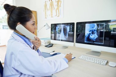 Asyalı genç kadın x-ray görüntüleri ile bilgisayarlara bakarak masada oturan ve ofiste telefonla konuşurken