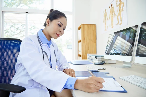 白いラボコートを着たアジアの若い女性医師がコンピュータの前のテーブルに座り オフィスのクリップボードに処方箋を書く — ストック写真