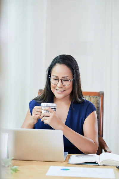 陽気なアジアのビジネスウーマンが朝のコーヒーを飲み 彼女のラップトップで電子メールをチェック — ストック写真