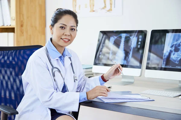 書類やX線画像で職場で働きながらカメラに微笑むアジアの若い女性放射線科医の肖像画 — ストック写真