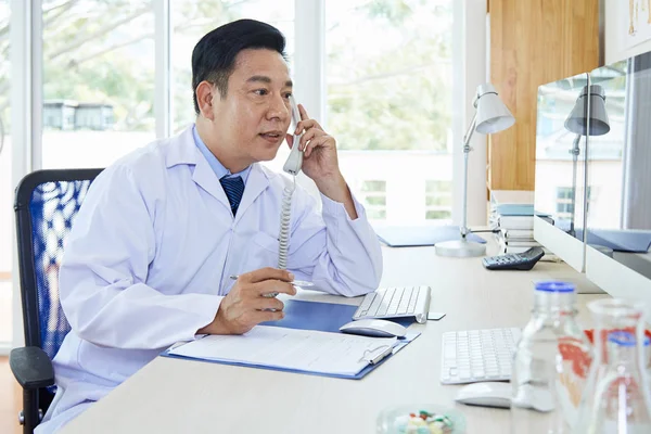 Reifer Asiatischer Arzt Laborkittel Sitzt Tisch Mit Klemmbrett Telefoniert Und — Stockfoto