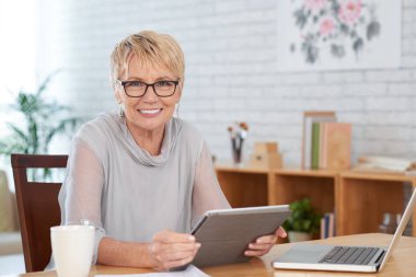 Oturma odasında masada oturan dijital tablet ile mutlu gülümseyen yaşlı kadın