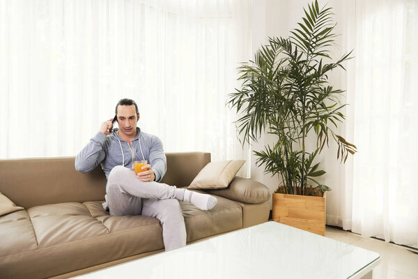 Serious man in comfortable clothes drinking fresh juice and calling on phone