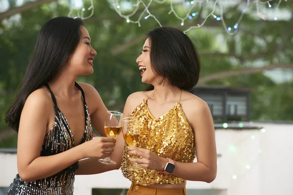 Laughing young Asian women in sparkling dresses drinking champagne and chatting at party