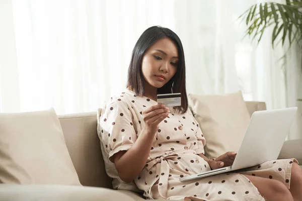 严肃的亚洲妇女坐在沙发上 用她的信用卡在家里用笔记本电脑在线支付 — 图库照片