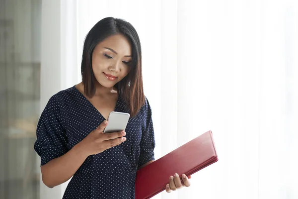 赤いフォルダを持つアジアの若い女性は 携帯電話でメッセージを入力したり 彼女のスマートフォンを使用してオンラインで作業 — ストック写真