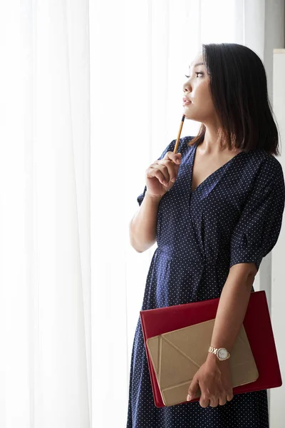 Asian businesswoman standing in beautiful dress with folder and digital tablet and thinking about new ideas while looking out of the window