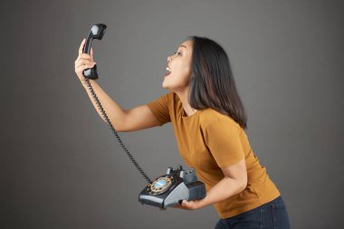 Gri arka planda dururken elinde retro telefon tutan ve cep telefonuyla bağıran genç bir kadın.