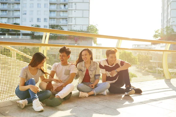 Happy Vietnamese young people in casual clothes sitting on bridge in rays of sun