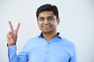 Barış işareti gösteren ve beyaz arka planda izole kamera gülümseyen mavi gömlekli Hintli genç işadamı portresi