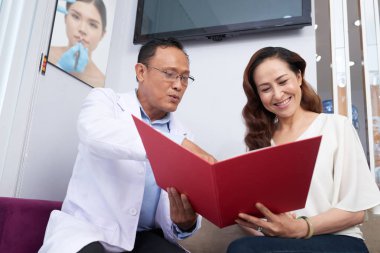 Asyalı olgun doktor kadın hasta ile birlikte kanepede oturan ve ofiste kırmızı klasörde onu farklı tıbbi prosedürleri gösteren 